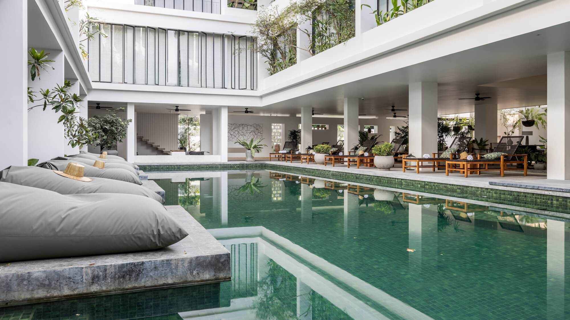 Modern indoor pool area with green-tiled water, lounge chairs, and seating surrounded by white columns and lush greenery. Natural light enhances this Siem Reap Luxury Accommodation’s open, tranquil design.