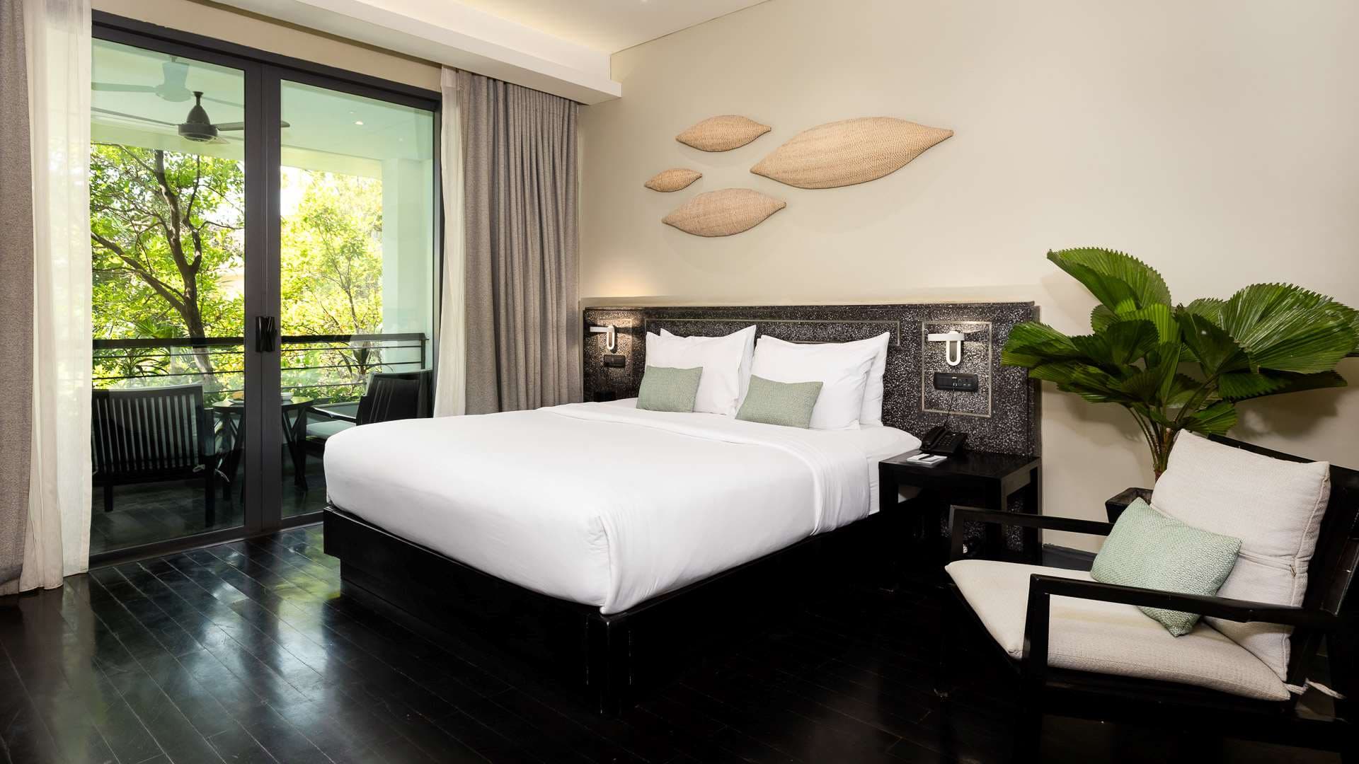 Anjali by Syphon modern hotel room in Siem Reap with a king-sized bed, white linens, dark wooden floor, and a balcony overlooking greenery. Decor includes minimalist wall art and a plant.