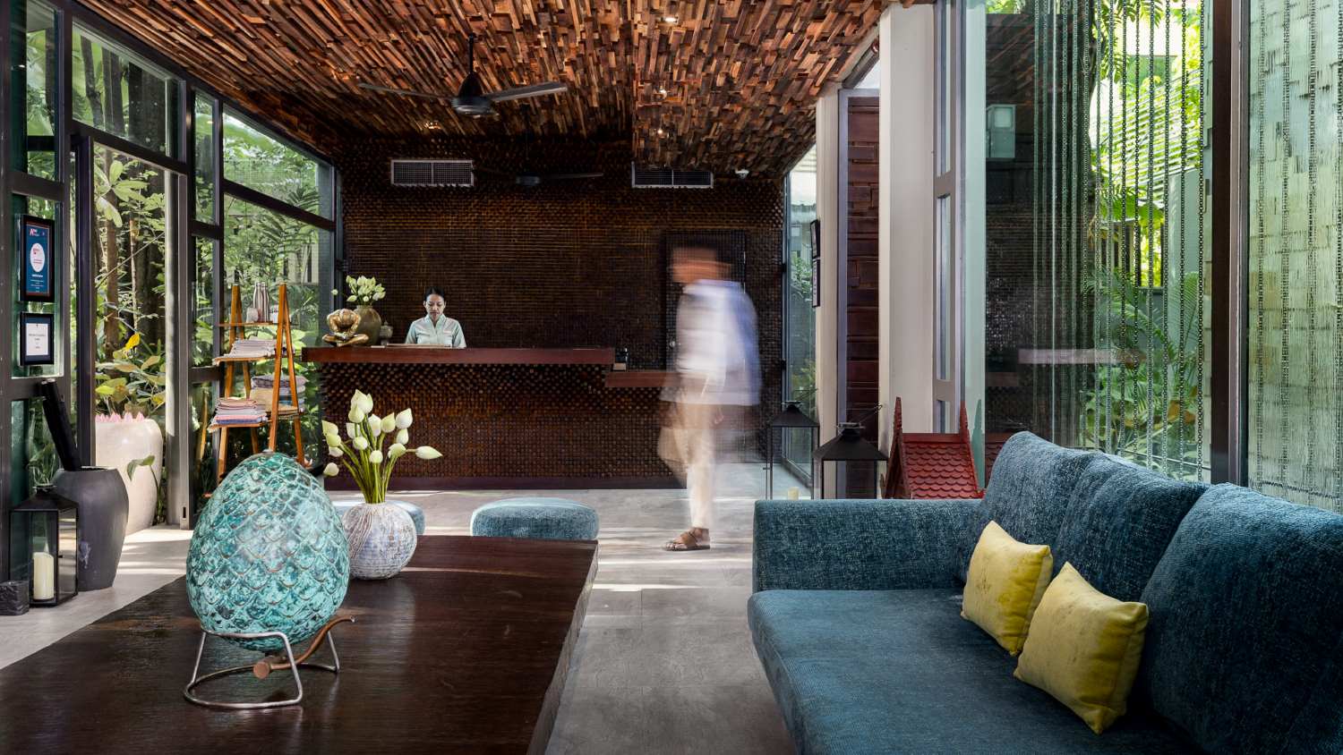 Modern hotel lobby in Anjali by Syphon, Siem Reap with a blurred person walking by, featuring a stylish reception desk, plush seating, and bamboo-inspired decor.